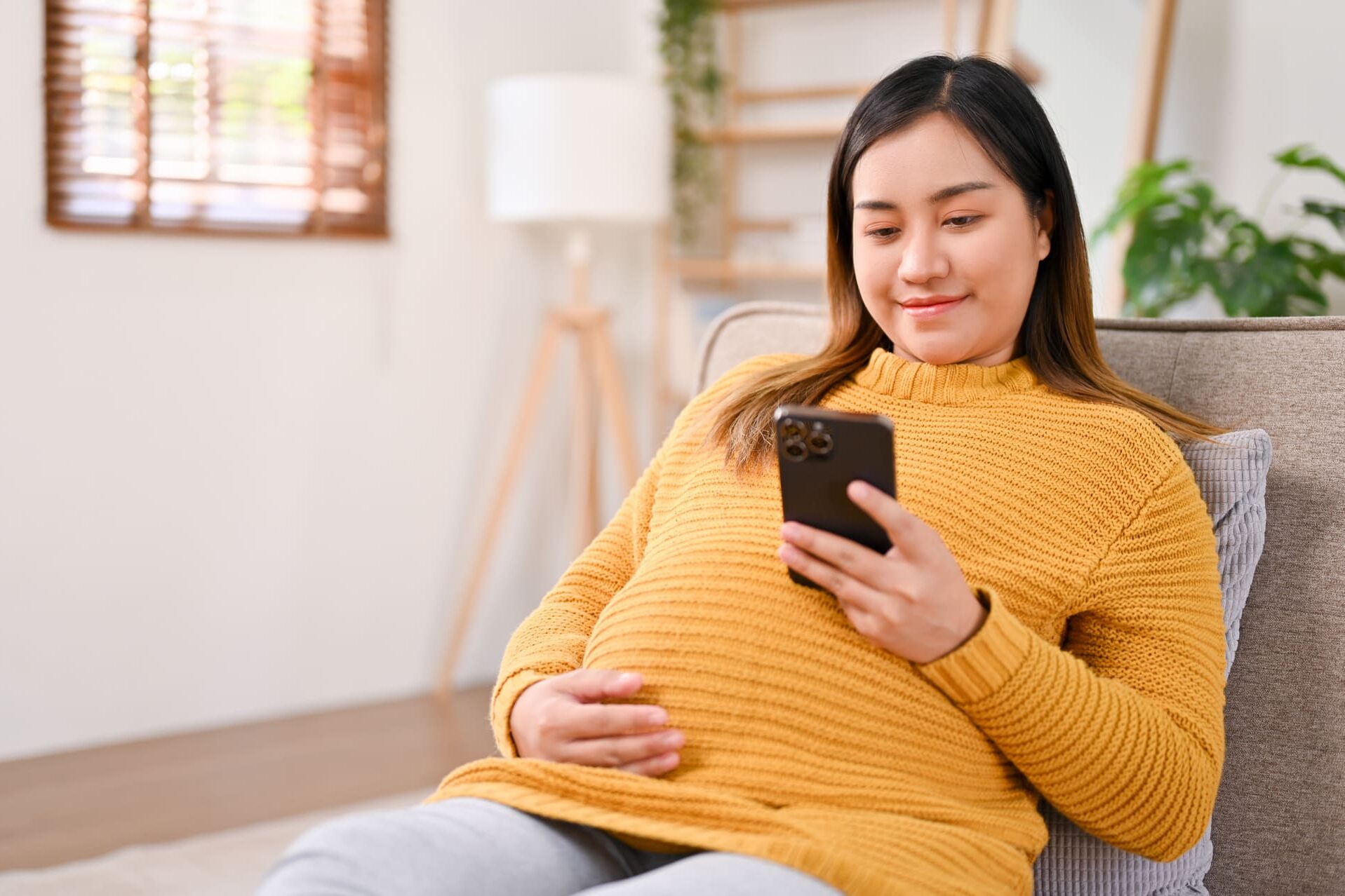 Relaxed Asian pregnant woman checking her baby's due date on her phone while resting on sofa 